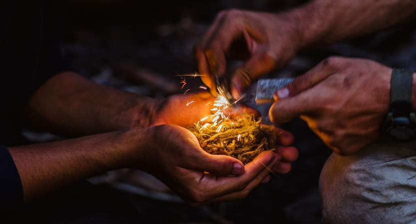 a person holds kindling in their hand while another works to light a spark 